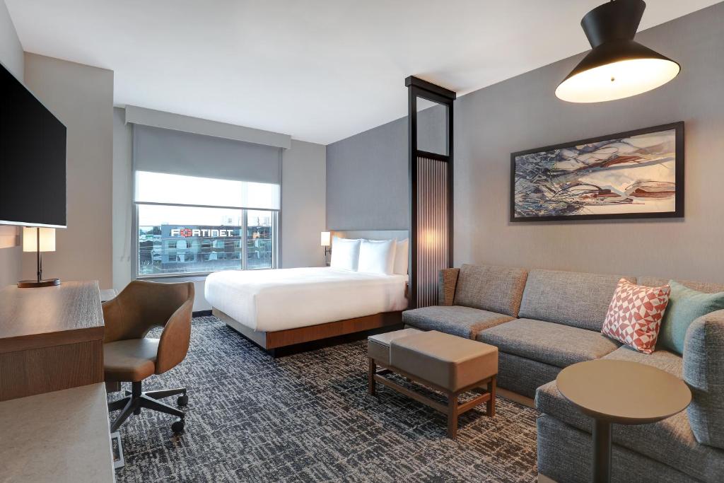 Hotel suite with a bed and a grey sofa, a coffee table, and a work desk near a large window overlooking the city outside the Fortinet sign, creating a modern, airy living space.