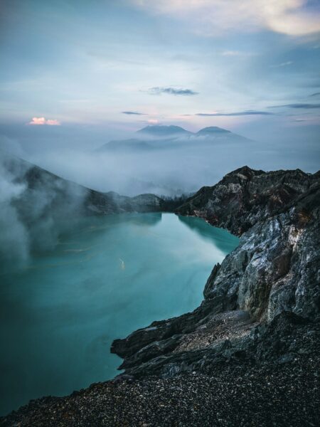 Turquoise crater lake edged by dark rocky cliffs and morning fog over distant mountains.