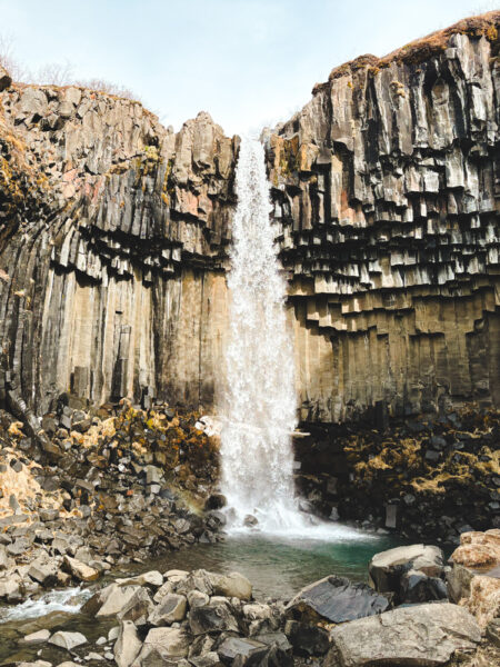 Waterfall cascading down basalt column cliffs into a rocky pool below.