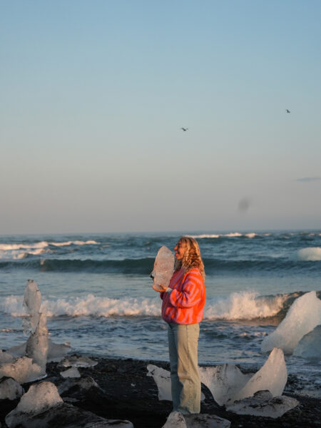 Woman in a bright pink-orange sweater holds a large piece of ice on a rocky beach with more ice chunks along the shore.
