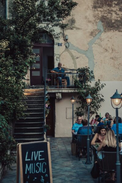 Man in a blue shirt on a balcony playing guitar above a busy outdoor cafe courtyard; chalkboard reads 'LIVE MUSIC'.