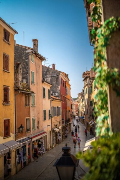 Sunlit European street with pastel buildings, shops, and people strolling along a narrow cobblestone road under a blue sky.