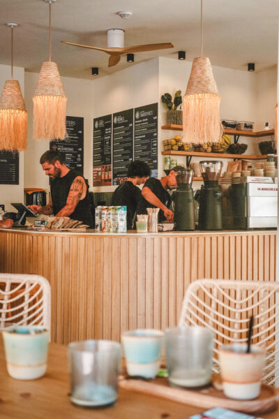 Cafe counter with baristas preparing drinks, chalkboard menus on the wall, and warm pendant lights above a wooden barrier.