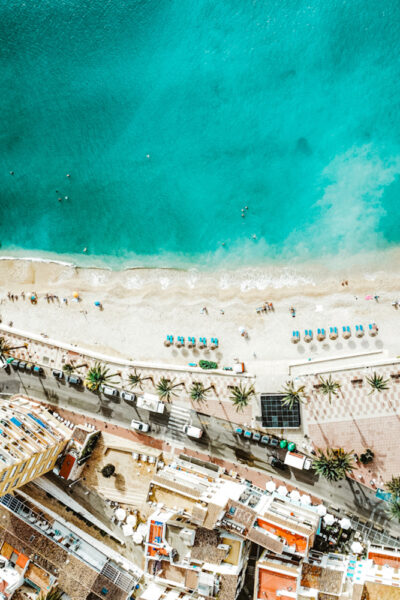 Aerial view of a turquoise sea meeting a sandy beach with rows of umbrellas and sunbathers along the shore, below a street lined with palm trees and buildings.