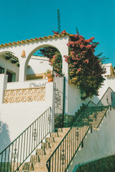 White stucco house with arched entry, stone stairs, black wrought-iron railing, and pink bougainvillea against a blue sky.