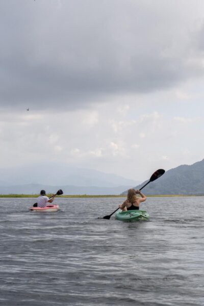 Two people kayaking on a large lake under gray clouds, with mountains in the background.