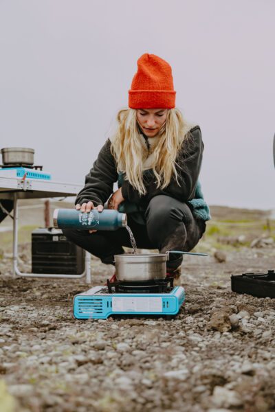 Woman in an orange beanie pours water into a pot on a blue camping stove outdoors by rocky ground.