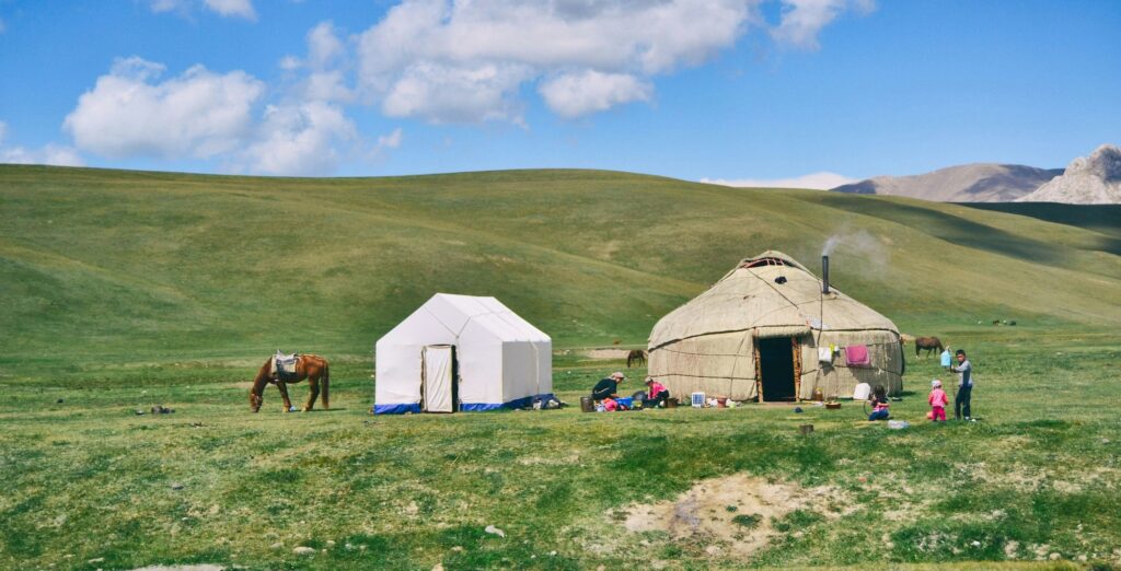 A campsite on a grassy plain with a white rectangular tent and a round yurt, people cooking nearby, children playing, and a horse grazing under a blue sky.