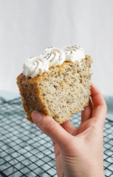 A hand holding a slice of vanilla cake with white whipped cream frosting and poppy seeds on top.