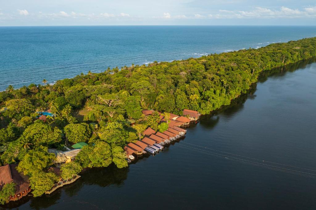 Aerial view of a tree-covered narrow land strip with the blue ocean on the left and a calm river on the right; brown-roofed cottages line the water’s edge.