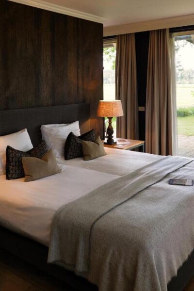 Hotel room with a king-size bed, plush pillows, and a lamp-lit wooden nightstand by a large window with beige curtains.
