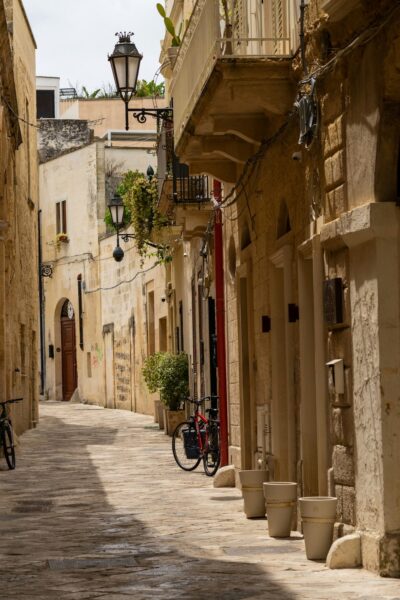 Narrow stone alley in a historic town with bicycles, potted plants, and warm sunlight.