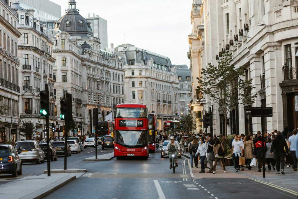 Busy city street in London with a red double-decker bus (route 94) and pedestrians along the sidewalk lined with historic buildings.