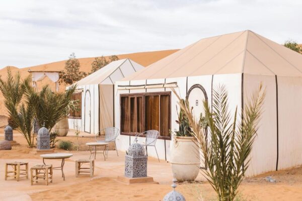 Desert glamping campsite with white canvas tents, beige roofs, palm plants, and metal lanterns on sand.