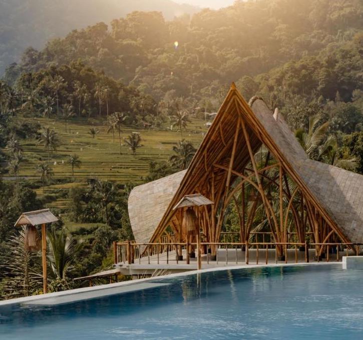 Wooden pavilion with a triangular roof beside an infinity pool, overlooking terraced green hills and palm trees at sunset/sunlight.