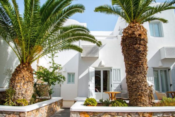 White Mediterranean villa with open double doors, stone planters, and tall palm trees on a sunny day.