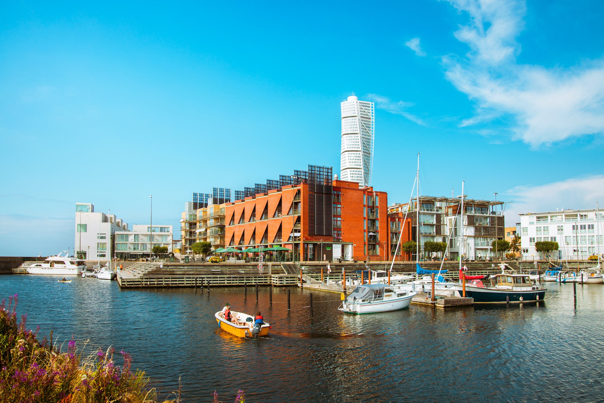 Harbor scene with colorful modern buildings along the water and several boats docked by a wooden quay under a bright blue sky.