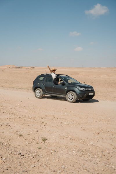 Person in a hat leaning out of a dark SUV, parked on a dusty desert road, raising a peace sign to the sky.