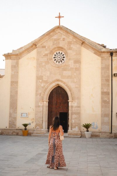 Woman in a patterned maxi dress approaches the arched wooden church door from the front, beige stone facade behind her.
