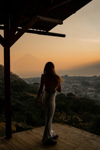 Woman in patterned leggings stands on a wooden deck at sunset, gazing over a valley toward a distant volcano on the horizon.