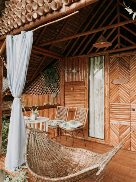 Bamboo veranda with a curved wicker hammock, a small white table with plants, two wooden chairs with leaf-pattern cushions, and a pale blue curtain.