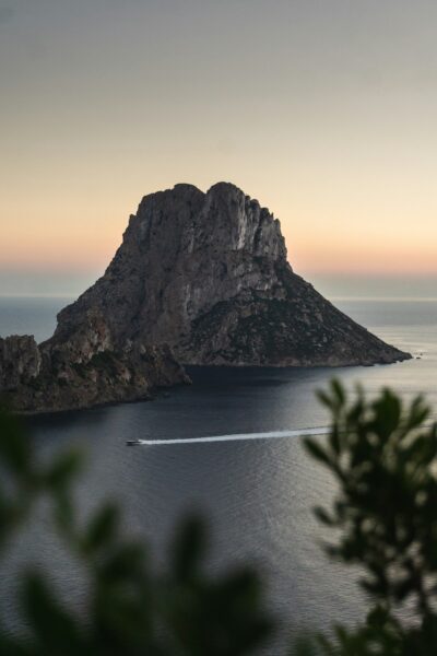 Rocky island peak rising from calm sea at sunset, with a small boat leaving a white wake nearby.