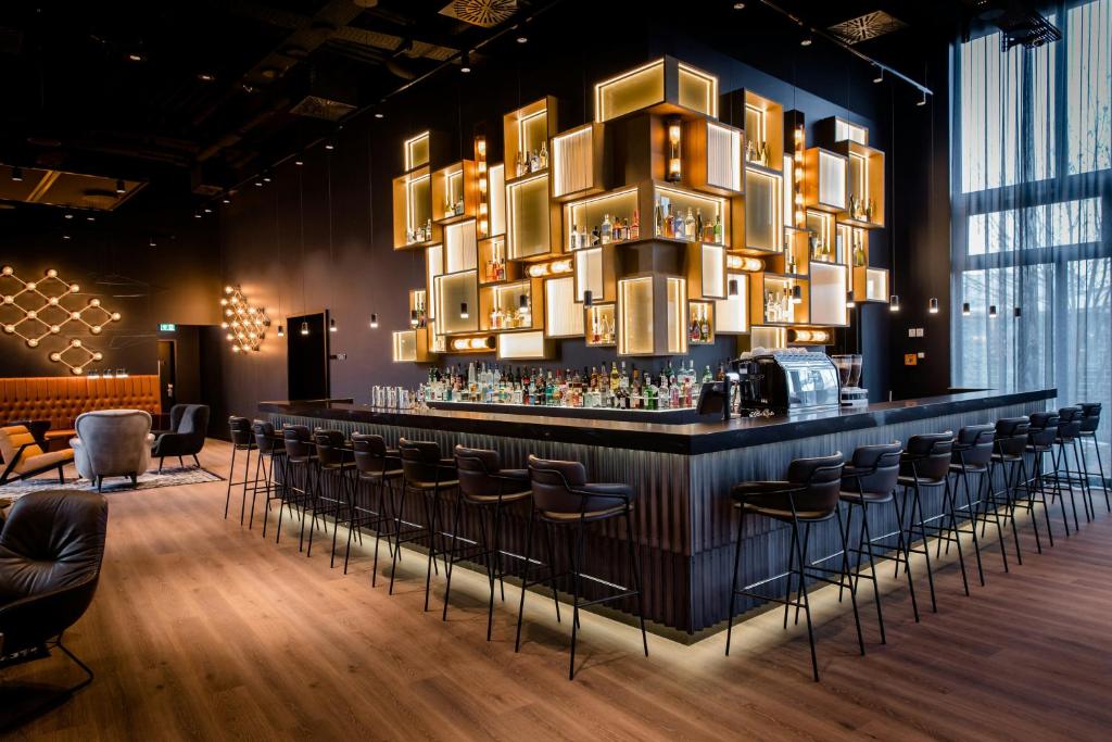 Stylish hotel bar with a long dark counter, black bar stools, and a geometric illuminated shelf display filled with bottles above the bar.