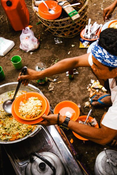 Man wearing a blue headband serves noodles from a pot into orange bowls at an outdoor street food setup with utensils nearby.]**Oops**