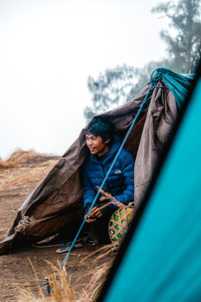 Young man in a blue puffer jacket inside a brown camping tent, handling a rope on a dirt campsite.