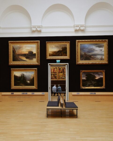 Museum gallery with five gold-framed landscape paintings on a dark wall, a wooden bench in the foreground, and two people sitting beyond a doorway.