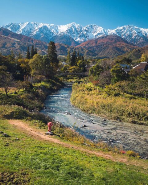 Winding river through a green valley with autumn trees and a distant range of snow-capped mountains under a clear blue sky; a pink lifebuoy statue sits by the riverbank.