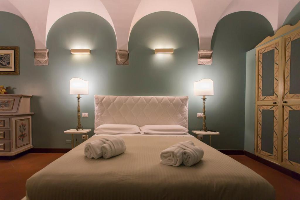 Hotel bedroom with teal walls and arched white ceiling, queen bed with tufted headboard, two bedside lamps, dresser left, ornate wardrobe right, and three rolled towels on the bed.