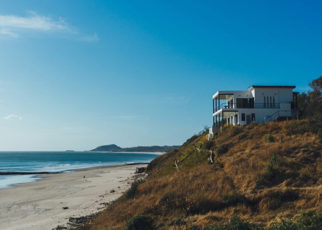 Modern white hillside house with a balcony overlooking a sandy beach and blue ocean under a clear sky.