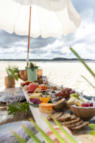 Beachside picnic spread on a low table under a white parasol, with fruit, cheese, bread, and glassware on the sand park by the sea at sunset.