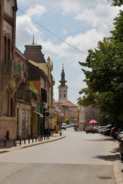 Urban street lined with colorful historic buildings and a distant church tower, with trees on the right and pedestrians visible.
