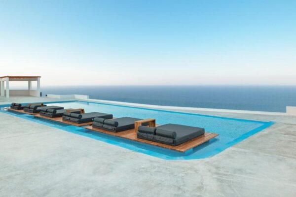 Infinity pool with black cushioned lounge chairs on wooden platforms, overlooking the ocean.