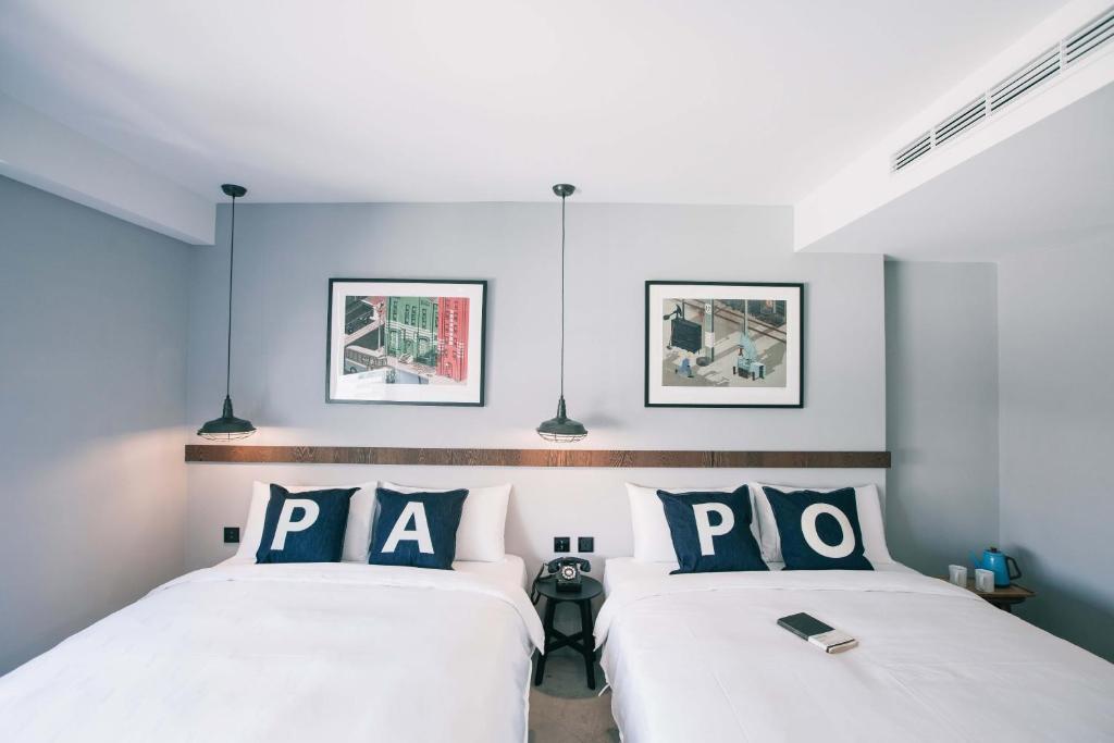 Twin hotel room with white bedding and navy pillows spelling 'PA' and 'PO' on each bed, a shared headboard, and two hanging pendant lights above.
