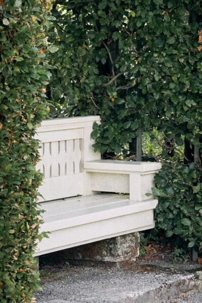 White wooden bench with a pale stone base tucked among dense green hedges in a sunny garden setting