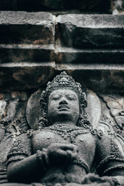 Stone statue of a deity with folded hands, ornate jewelry, weathered surface against a stone wall area.