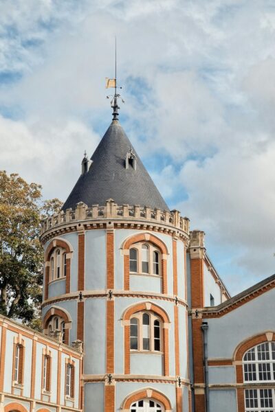 Round turreted section of a blue-and-brick historic building with arched windows and a tall cone-shaped slate roof topped by a weather vane, against a cloudy sky.