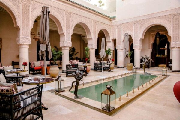 Moroccan-style courtyard with ornate arches surrounding a rectangular shallow pool and outdoor dining tables around it, with umbrellas and lanterns nearby.