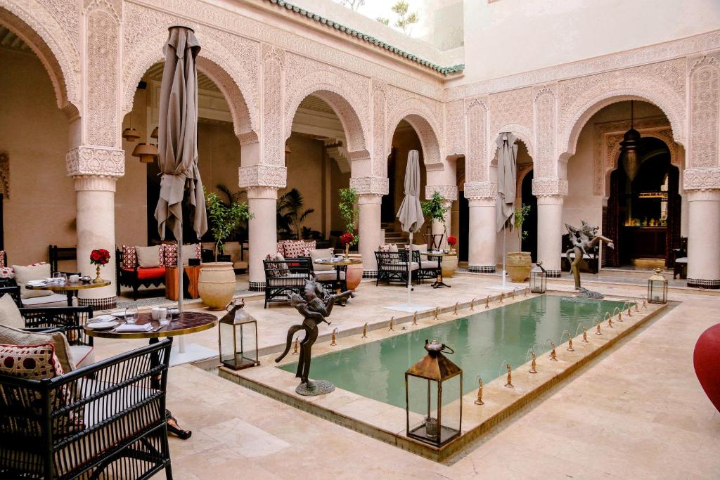 Moroccan-style courtyard with ornate arches surrounding a rectangular shallow pool and outdoor dining tables around it, with umbrellas and lanterns nearby.