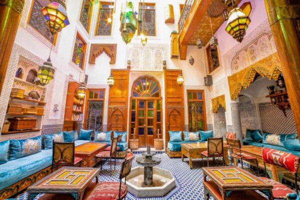 Bright Moroccan courtyard lounge with turquoise couches, colorful cushions, tiled floor, and a central fountain beneath hanging lanterns.