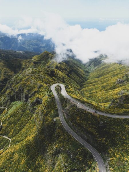 Winding mountain road cuts through green hills dotted with yellow flowers, under a cloudy sky viewed from above.