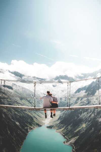 Two people sit close together on a narrow suspension bridge high above a turquoise alpine lake, surrounded by snow-capped mountains.