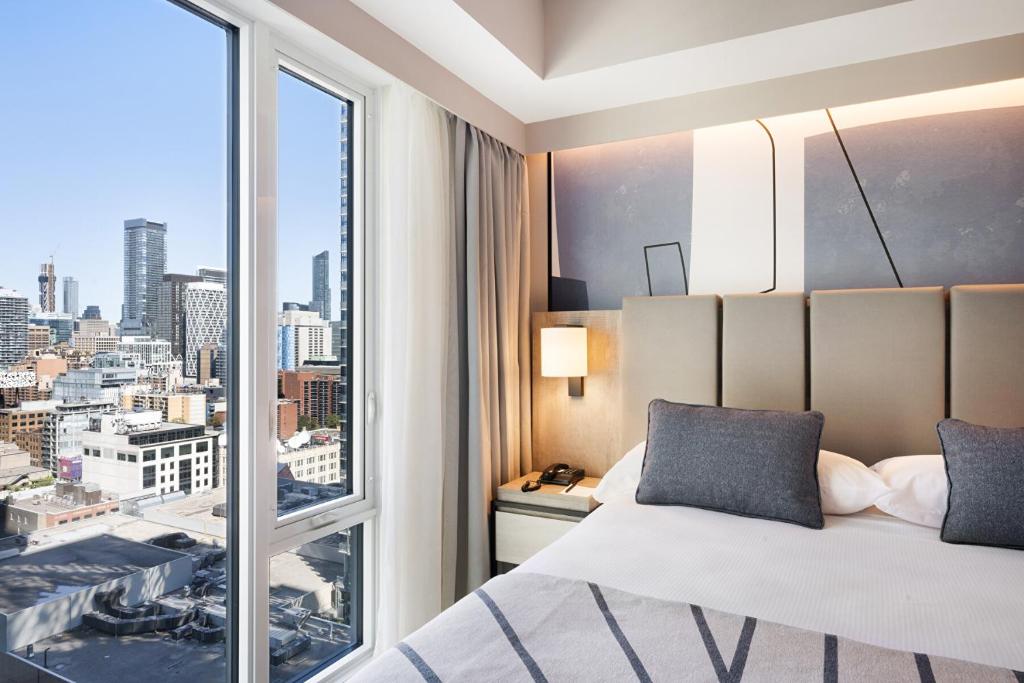Hotel room with a large window overlooking a city skyline, bed with gray pillows, and a bedside lamp.