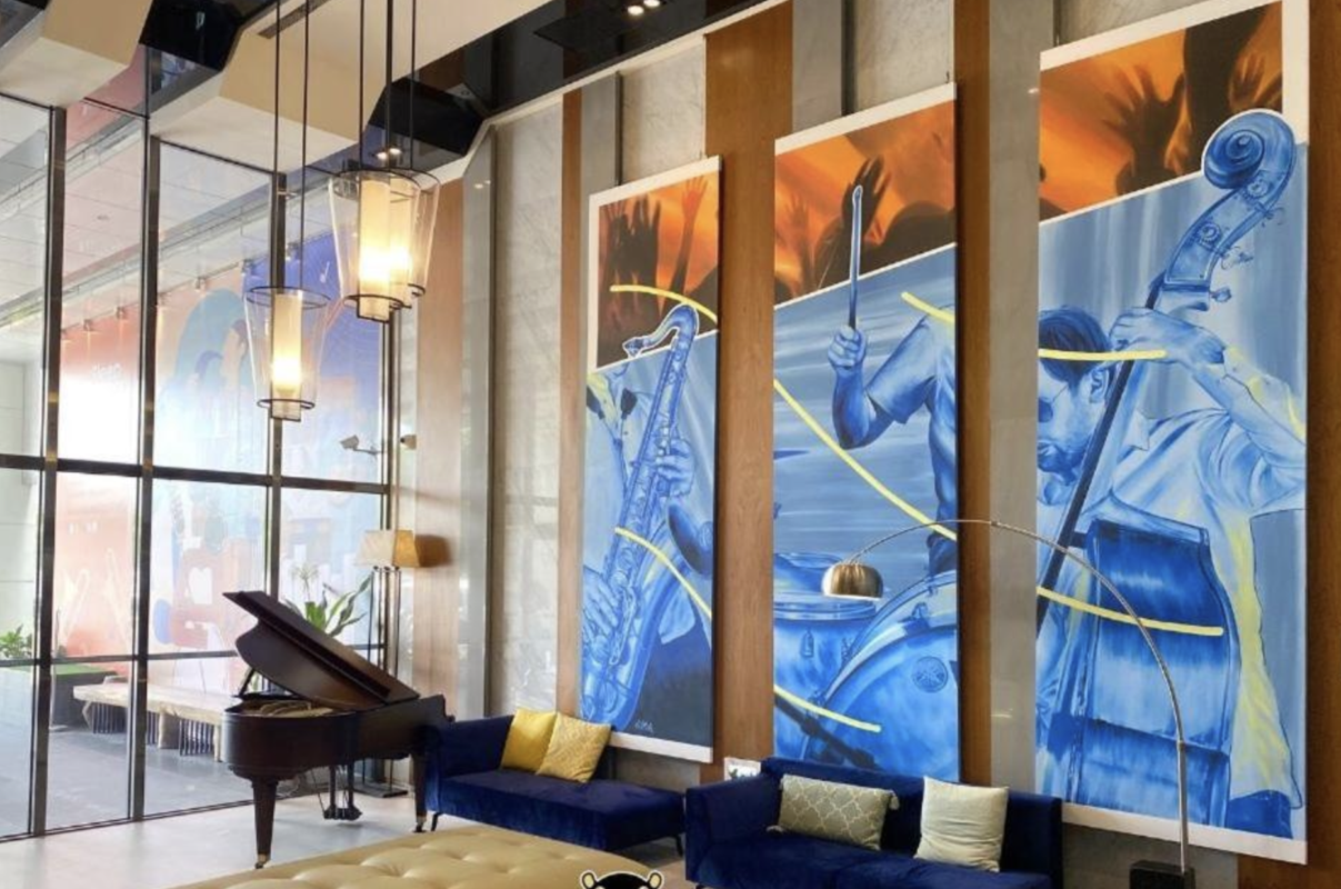 Contemporary hotel lobby with three large blue mural panels of musicians, a grand piano, glass walls, and a blue seating area with yellow cushions.