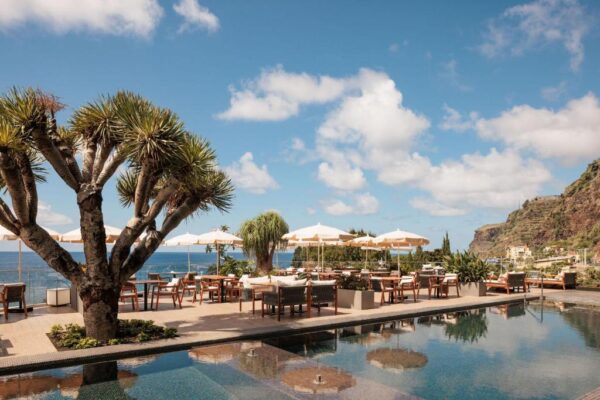 Poolside resort terrace with wooden chairs and beige umbrellas overlooking the sea and rocky coastline.
