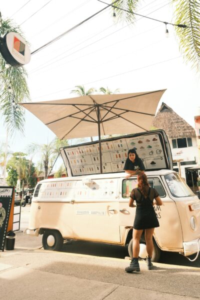 Cream-colored vintage food truck with a large menu board, beige umbrella, and a woman standing nearby on a sunny street. cool tropical setting with palm trees above.