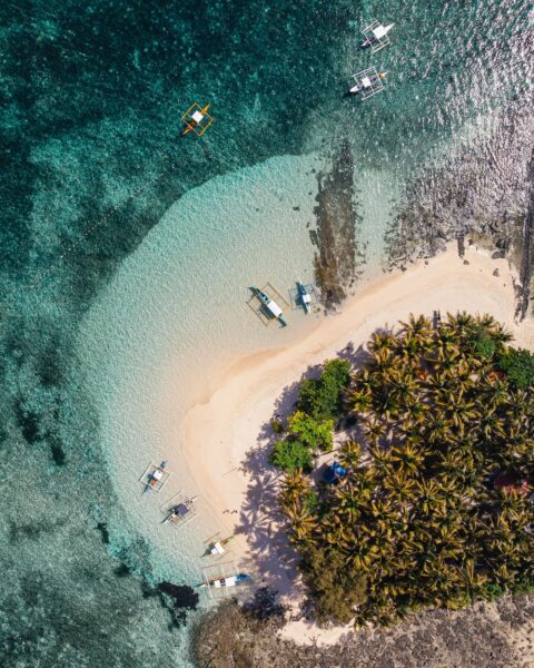 Aerial view of a tropical island with palm trees, white sand beach, and several small boats anchored near the shore.